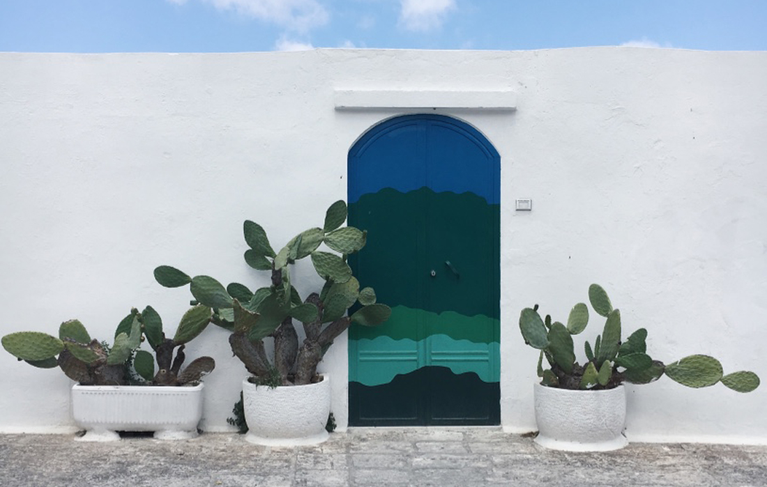 La porta azzurra di Ostuni, una porta pitturata sui toni dell'azzurro su una costruzione bianca. Ai lati, due cactus e sopra il cielo azzurro.