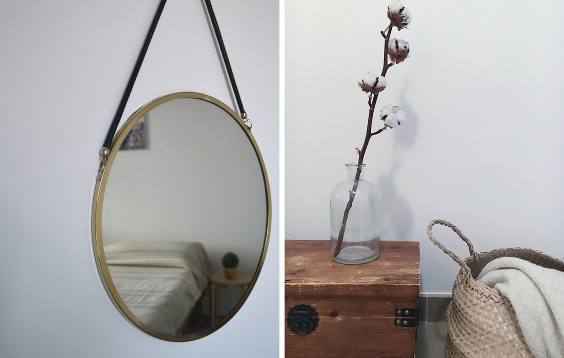 Dettagli di arredo scandinavo: uno specchio rotondo con cornice bronzo e una vecchia cassetta in legno con sopra un vaso con un fiore di cotone. Accanto una cesta di paglia dalla quale esce una coperta panna.