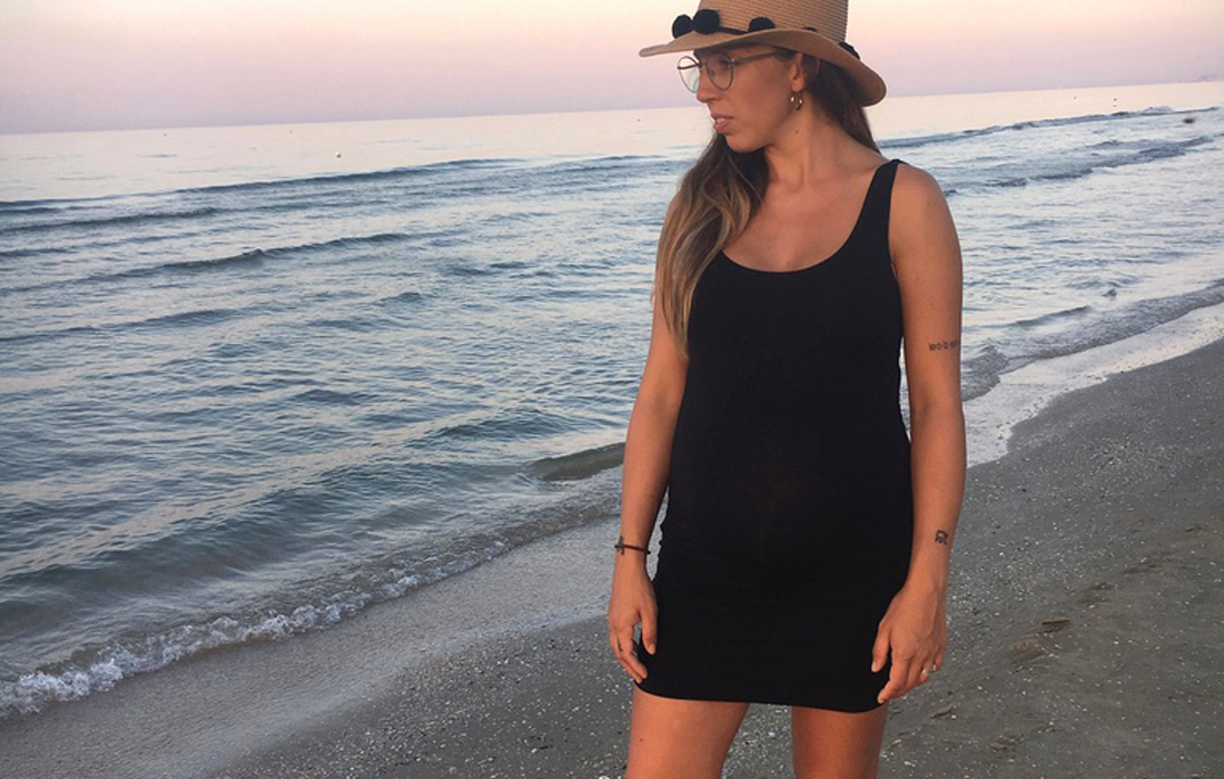 Donna incinta all'ottavo mese di gravidanza sulla riva del mare con abito corto nero e cappello in paglia, prospettiva frontale