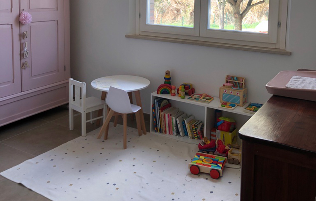 Cameretta da bambina con mobile basso, giochi in legno. Un tappeto a triangoli colorati e un tavolo basso rotondo bianco con gambe in legno naturale e due seggioline basse