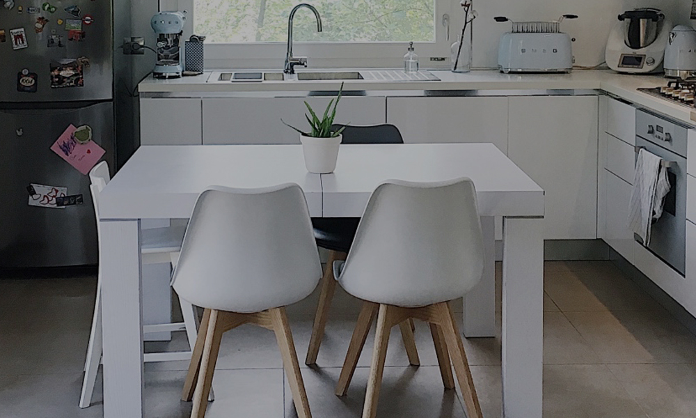 Una cucina in stile scandinavo con tavolo in legno bianco e sedie con zampe in legno chiaro. Sullo sfondo un frigo in acciaio e una grande finestra