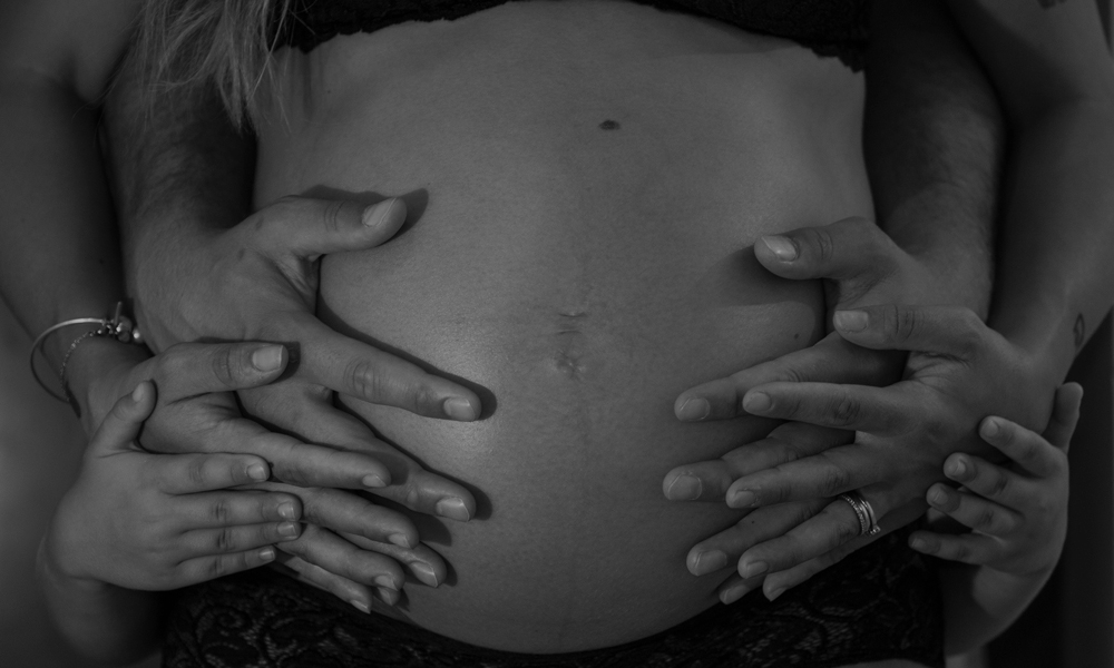 In primo piano un pancione di una donna in gravidanza con appoggiate le mani della mamma, del babbo e del fratellino