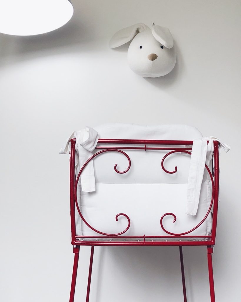 Culla in ferro battuto rossa con rivestimento bianco. Appesa al muro una decorazione a forma di testa di coniglio in stoffa. In alto a sinistra una lampada