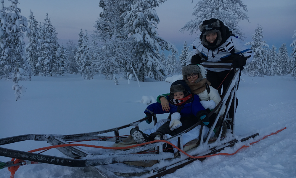 Un uomo, una donna e un bambino su una slitta trainata dagli husky. Sullo sfondo la foresta lappone.