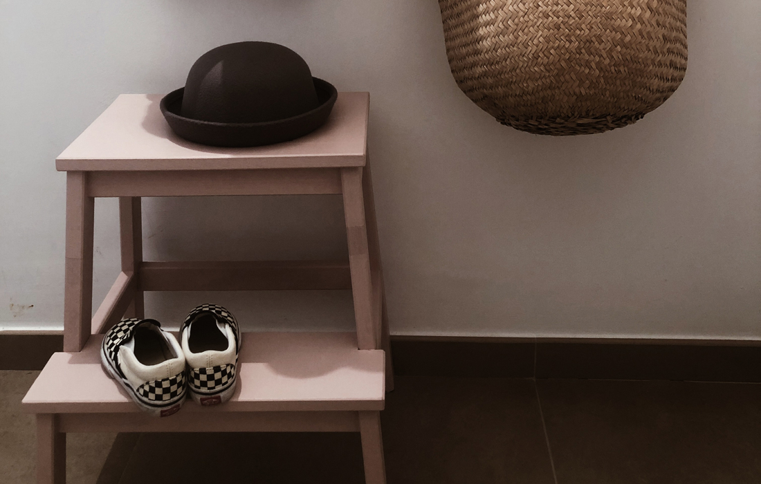 Sgabello basso per bambini con scarpe appoggiate sopra e un cappello. Di lato, una cesta in vimini per riporre cappelli, sciarpe e altri accessori.